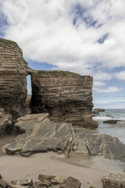Katmanlı kaya oluşumları ve doğal kemerleriyle dramatik sahil kayalıkları parçalı bulutlu bir gökyüzünün altında denizle buluşuyor.