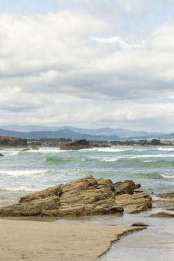 Kıyı manzarası, kayalık çıkıntılar ve turkuaz dalgalar kumlu bir plaja yuvarlanıyor ve dağlar uzaklardan görünüyor.