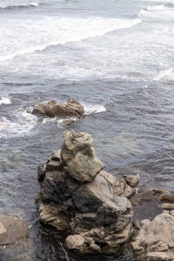 Sivri kıyı kayaları sığ deniz sularından yükselir, parlak, bulutlu bir gökyüzünün altında, yumuşak okyanus dalgalarıyla çevrilidir.