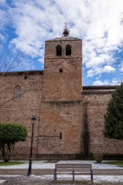 Yıpranmış bir duvarın üzerinde yükselen taş kilise kulesi, ön planda bir ahşap bank ve bir sokak lambası var, bulutlu bir gökyüzünün altında.