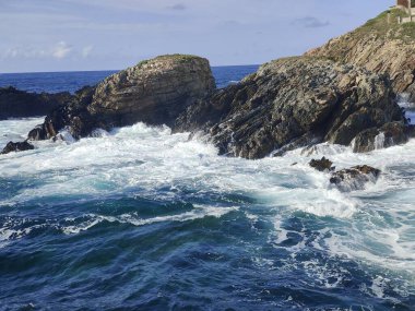 Güçlü okyanus dalgaları kayalık bir kıyı şeridine çarpıyor, parlak güneşli bir günde beyaz köpük ve sprey yaratıyor.