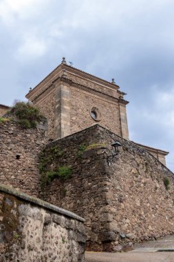 İspanya, Candelario 'daki yıpranmış bir duvarın üzerinde yükselen tarihi taş kilise kulesinin alçak açılı görüntüsü bulutlu bir gökyüzünün altında.