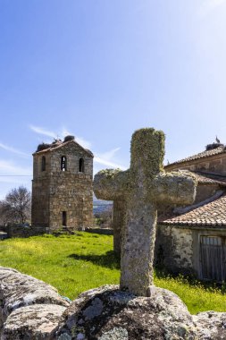 Güneşli bir günde çatıda leylek yuvaları olan eski bir kilisenin önünde duran yosunlu taş haç.