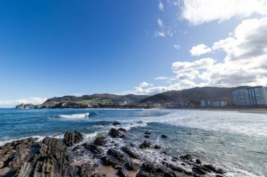 Dalgaların kayalık bir kıyı şeridine çarpması manzarası, Bakio şehrinin manzarası, İspanya, uzaktaki tepelerin arasına yerleşmiş.