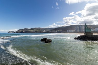 Okyanus dalgaları Bakio 'daki sahile doğru akıyor, Bask bölgesi, İspanya, arkasında binalar ve dağlar var.