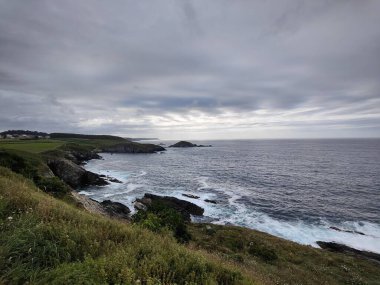 Dalgalar dramatik bulutlu bir gökyüzü altında kayalık bir sahile çarpıyor. Güzel ve güçlü bir deniz manzarası yaratıyor.