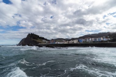 Okyanus dalgaları bulutlu bir gökyüzünün altında kıyı kasabası olan kayalık bir kıyı şeridine çarpıyor.