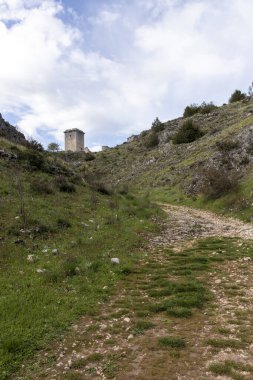 Bulutlu mavi gökyüzünün altındaki tarihi gözetleme kulesine doğru giden kayalık yol İspanya kırsalında macera ve keşif hissi uyandırıyor.