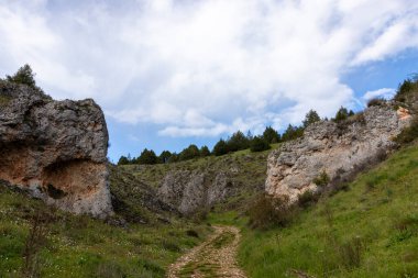 Bulutlu bir gökyüzünün altında, büyük kaya oluşumları ve yeşil tepeler arasında geçen manzaralı yürüyüş patikaları.
