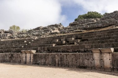 Bulutlu bir gökyüzünün altındaki eski bir amfi tiyatroda yıpranmış taşlar.