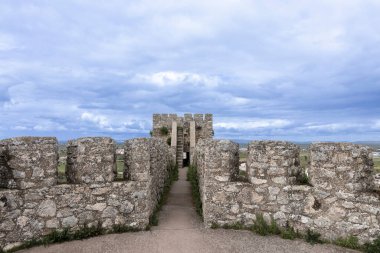 Antik taş duvarlar arasındaki yol bir gözetleme kulesine çıkıyor, bulutlu bir gökyüzü dramatik bir zemin oluşturuyor.