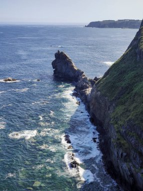 Engebeli bir kıyı şeridi manzarası belirgin bir deniz yığını, kayalara çarpan dalgalar ve bitki örtüsüyle kaplı kayalıklar.