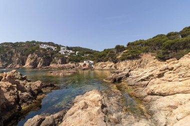 Huzurlu turkuaz sular kayalık bir kıyı şeridine nazikçe sarılır, İspanya 'da berrak mavi bir gökyüzünün altında kayalıkların kenarındaki evleri beyazlatır.