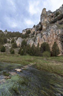 Arkaplanda bulutlu bir gökyüzünün altında yükselen dik kayalık bir uçurum ile yeşil bir çayırda kıvrılan ucero nehrinin manzarası