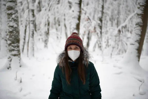 a woman standing in the middle of a snow-covered forest, wearing a white mask, teal winter jacket, and a multicolored hat. The trees around her are draped in snow, emphasizing the quiet of the winter season