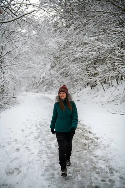 The photograph shows a woman walking along a snow-covered path in the forest. She is dressed in a teal jacket and a colorful knitted hat, with snow-laden trees forming a natural archway over the trail.