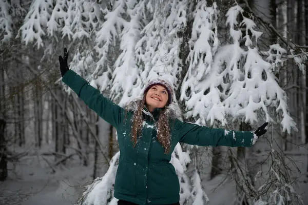 The image depicts a woman in a snow-filled forest, arms joyfully raised. She is wearing a teal winter jacket and a snow-speckled knitted hat, with a backdrop of heavily snow-laden branches