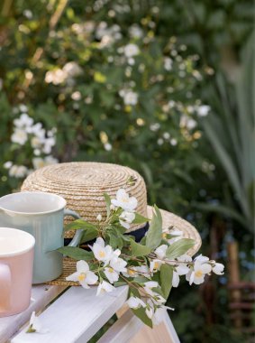 Yasemin dallı bir hasır şapka ve bahçedeki beyaz ahşap masada iki bardak. Yasemin çiçekli bir çalılığın arka planına karşı. Doğada rahatla.