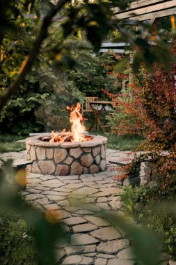 Patio and fire in the fire pit in the evening. Beautiful fire flames surrounded by beautiful trees. In the background is a table and two chairs. Summer collection clipart