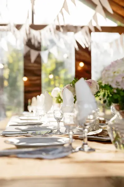 A long festive table decorated with white flowers and greenery; there are plates, glasses and candles on the table. The room is decorated with white garlands. Wedding decorations