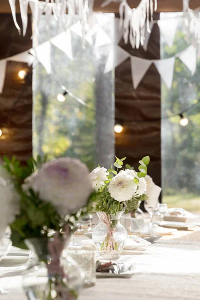 A long festive table decorated with white flowers and greenery; there are plates, glasses and candles on the table. The room is decorated with white garlands. Wedding decorations