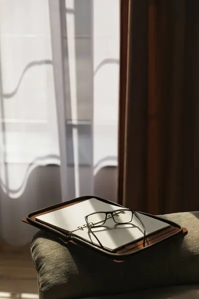 Glasses and an open weekly book lay on a couch in a room with dimmed light. There is a beautiful pattern of light and shadow on the furniture and on the diary. Vertical image.