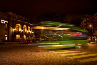A green city bus drives down a night street in the light of street lamps past the Landskrona Theatre. Long exposure. Blurred lights of a traveling tarnsport at speed. Landskrona Sweden 14.10.2022
