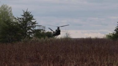 Çift kanatlı helikopter havalanıyor ve ağaçların arkasında kayboluyor.