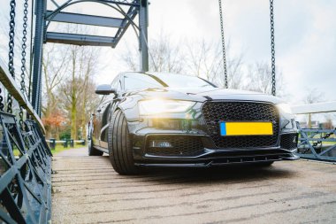 Görüntü, açık havada metal bir köprüye park etmiş koyu renkli bir sedan araba gösteriyor..