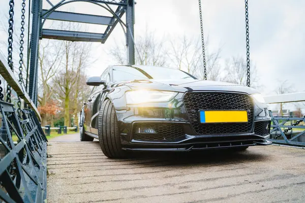 Görüntü, açık havada metal bir köprüye park etmiş koyu renkli bir sedan araba gösteriyor..