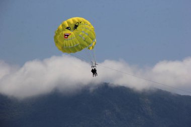 Budva plajı yakınlarında mavi bir gökyüzünde paraşüt, insanlarla dolu sarı bir paraşüt.
