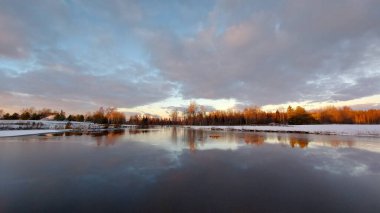 Kış sahnesinde suda yansımalar ve ağaçta günbatımı rengi. Kış boyunca mavi bulutlu gökyüzüne karşı donmuş ağaçlar.
