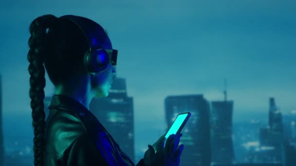 メガネやヘッドフォンでスマートフォンとサイバーパンクの女の子の肖像画 都市の高層ビルの背景に美しい若い女性 未来的概念 — ストック動画