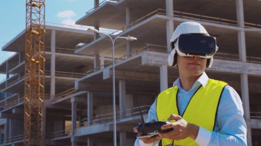 Sanal gerçeklik kasklı profesyonel dron operatörü inşaat alanının önünde duruyor. Uzaktan kumandalı bir bina. Ofis binası ve vinç arka planı. İş, gayrimenkul ve