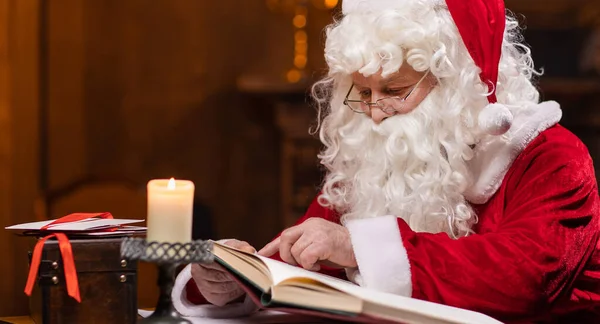 Workplace of Santa Claus. Cheerful Santa is reading the book of wishes while sitting at the table. Fireplace and Christmas Tree in the background. Traditional Christmas concept.