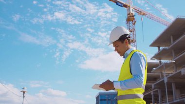 İnşaat alanının önünde duran profesyonel müteahhit. Kasklı ve yelekli ustabaşı. Ofis binası ve vinç arka planı. İş, gayrimenkul ve yatırım kavramları.