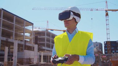 Sanal gerçeklik kasklı profesyonel dron operatörü inşaat alanının önünde duruyor. Uzaktan kumandalı bir bina. Ofis binası ve vinç arka planı. İş, gayrimenkul ve