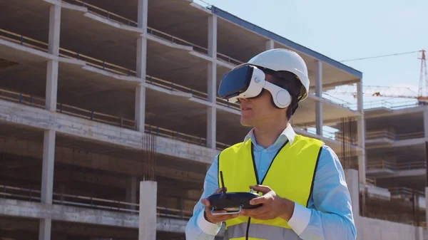 Professional drone operator in virtual reality helmet standing in front of construction site. Builder holding remote controller. Office building and crane background. Business, real estate and