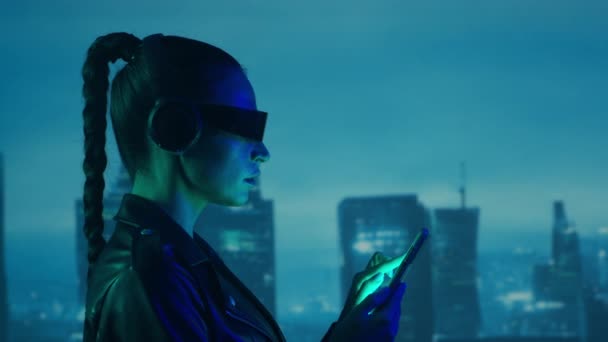 メガネやヘッドフォンでスマートフォンとサイバーパンクの女の子の肖像画 都市の高層ビルの背景に美しい若い女性 未来的概念 — ストック動画
