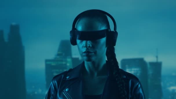 眼鏡とヘッドフォンのサイバーパンクの女の子の肖像画 都市の高層ビルの背景に美しい若い女性 未来的概念 — ストック動画