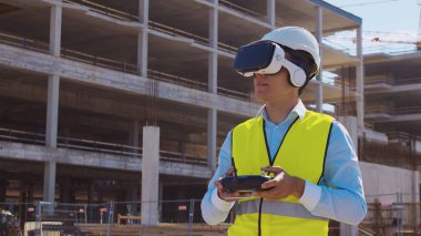 Sanal gerçeklik kasklı profesyonel dron operatörü inşaat alanının önünde duruyor. Uzaktan kumandalı bir bina. Ofis binası ve vinç arka planı. İş, gayrimenkul ve