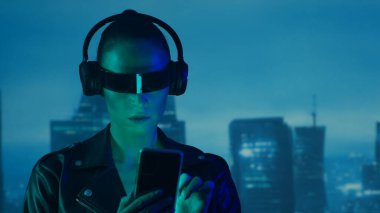 Gözlüklü ve kulaklıklı akıllı telefonlu siber punk kız portresi. Şehir tırpanlarının arka planında güzel bir genç kadın. Gelecekçi kavram.