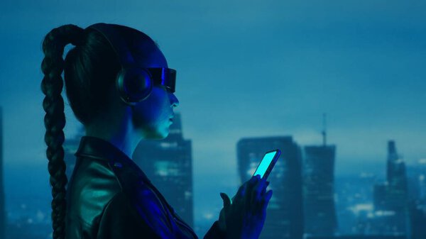 Portrait of cyberpunk girl with a smartphone in a glasses and headphones. Beautiful young woman on the background of city scyscrapers. Futuristic concept.