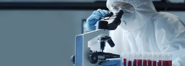 Scientist in protection suit and masks working in research lab using laboratory equipment: microscopes, test tubes. Coronavirus 2019-ncov hazard, pharmaceutical discovery, bacteriology and virology