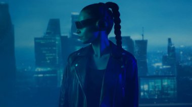 Gözlüklü ve kulaklıklı akıllı telefonlu siber punk kız portresi. Şehir tırpanlarının arka planında güzel bir genç kadın. Gelecekçi kavram.