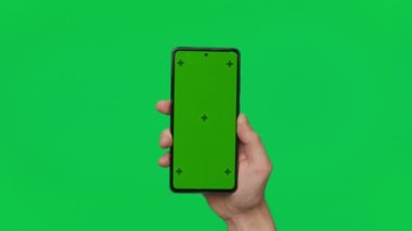 Male hand holding a smartphone with vertical green chroma key screen isolated on green background. Mobile phone in a hand. Different signs and gestures with fingers. 4K template. Technology and
