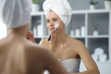 Young woman sits in the bathroom in front of the makeup mirror and does cosmetic procedures. Beautiful girl in white towel. The concept of skin care, health, rejuvenation and spa treatment.