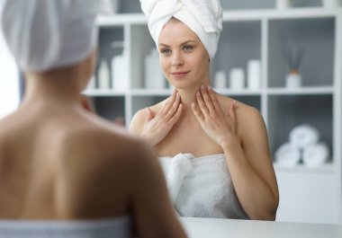 Young woman sits in the bathroom in front of the makeup mirror and does cosmetic procedures. Beautiful girl in white towel. The concept of skin care, health, rejuvenation and spa treatment.