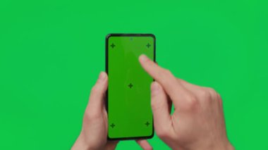 Male hand holding a smartphone with vertical green chroma key screen isolated on green background. Mobile phone in a hand. Different signs and gestures with fingers. 4K template. Technology and