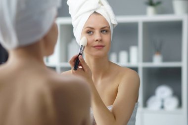 Young woman sits in the bathroom in front of the makeup mirror and does cosmetic procedures. Beautiful girl in white towel. The concept of skin care, health, rejuvenation and spa treatment.